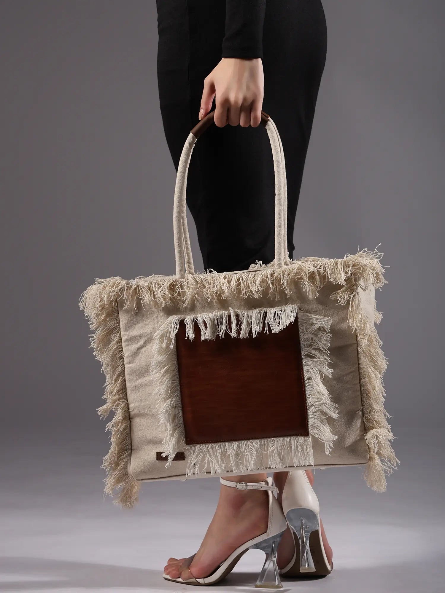 Person holding a beige tote bag with fringe details against a gray background By ENSAC