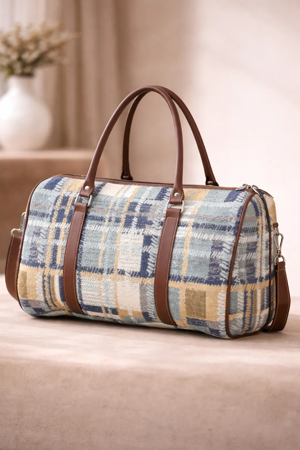 Plaid patterned duffel bag with brown leather straps on a beige surface BY ENSAC.